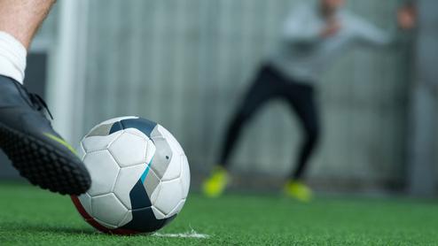 Nahaufnahme eines Fußballes, der auf grünem Kunstrasen vor einem verschwommenen Spieler liegt.