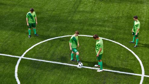 Vier Fußballspieler in grünen Trikots stehen auf einem Kunstrasenfeld, während ein Ball in der Mitte wartet.